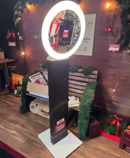Fotomatón con aro de luz LED instalado en una zona decorada con temática navideña. El dispositivo muestra una pantalla con la interfaz de toma de fotos, rodeado de elementos decorativos como guirnaldas, cajas de regalo, un banco de madera y luces cálidas.