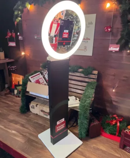 Fotomatón con aro de luz LED instalado en una zona decorada con temática navideña. El dispositivo muestra una pantalla con la interfaz de toma de fotos, rodeado de elementos decorativos como guirnaldas, cajas de regalo, un banco de madera y luces cálidas.
