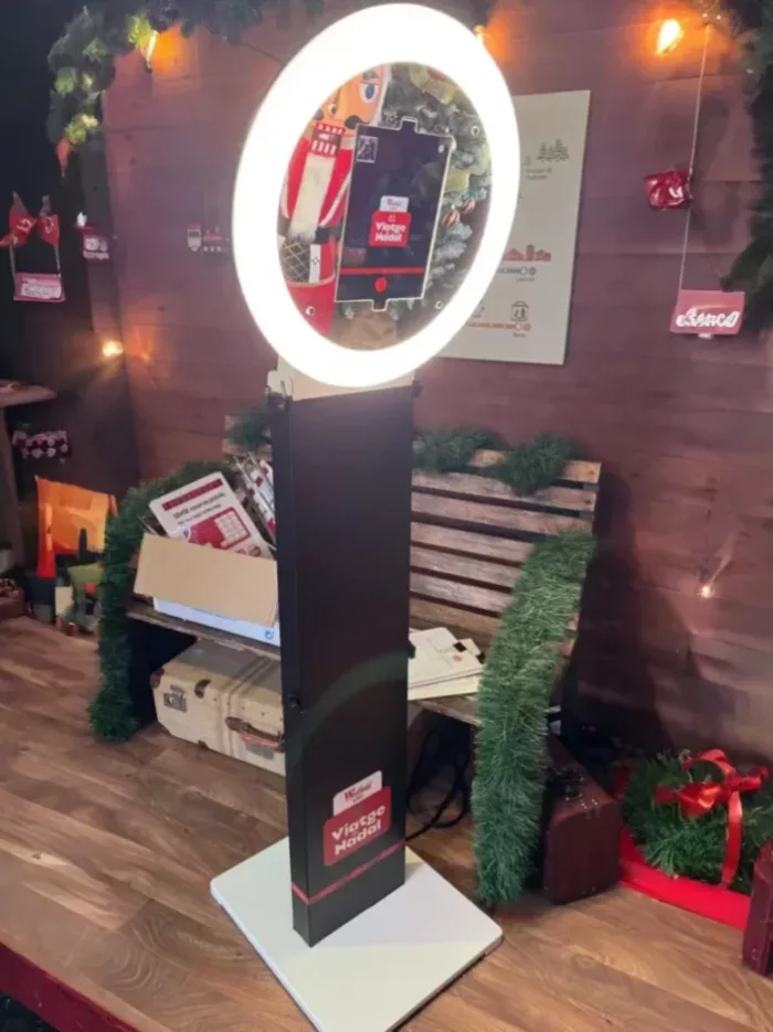 Fotomatón con aro de luz LED instalado en una zona decorada con temática navideña. El dispositivo muestra una pantalla con la interfaz de toma de fotos, rodeado de elementos decorativos como guirnaldas, cajas de regalo, un banco de madera y luces cálidas.