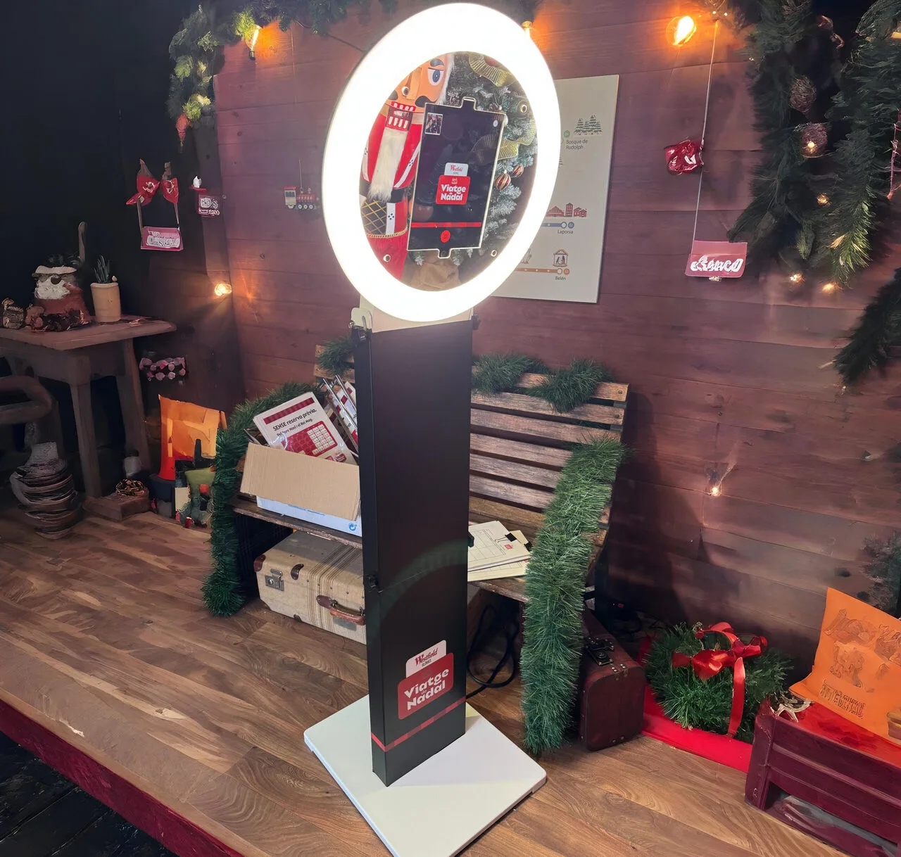 Fotomatón con aro de luz LED instalado en una zona decorada con temática navideña. El dispositivo muestra una pantalla con la interfaz de toma de fotos, rodeado de elementos decorativos como guirnaldas, cajas de regalo, un banco de madera y luces cálidas.